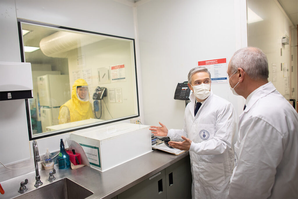 Meric Gertler and François-Philippe Champagne both wear masks and lab coats. They look through a window into secure lab.