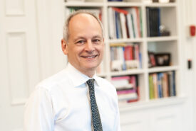 U of T President Meric Gertler smiles, leaning on a desk. Behind him, a cabinet-style bookshelf full of books and pictures.